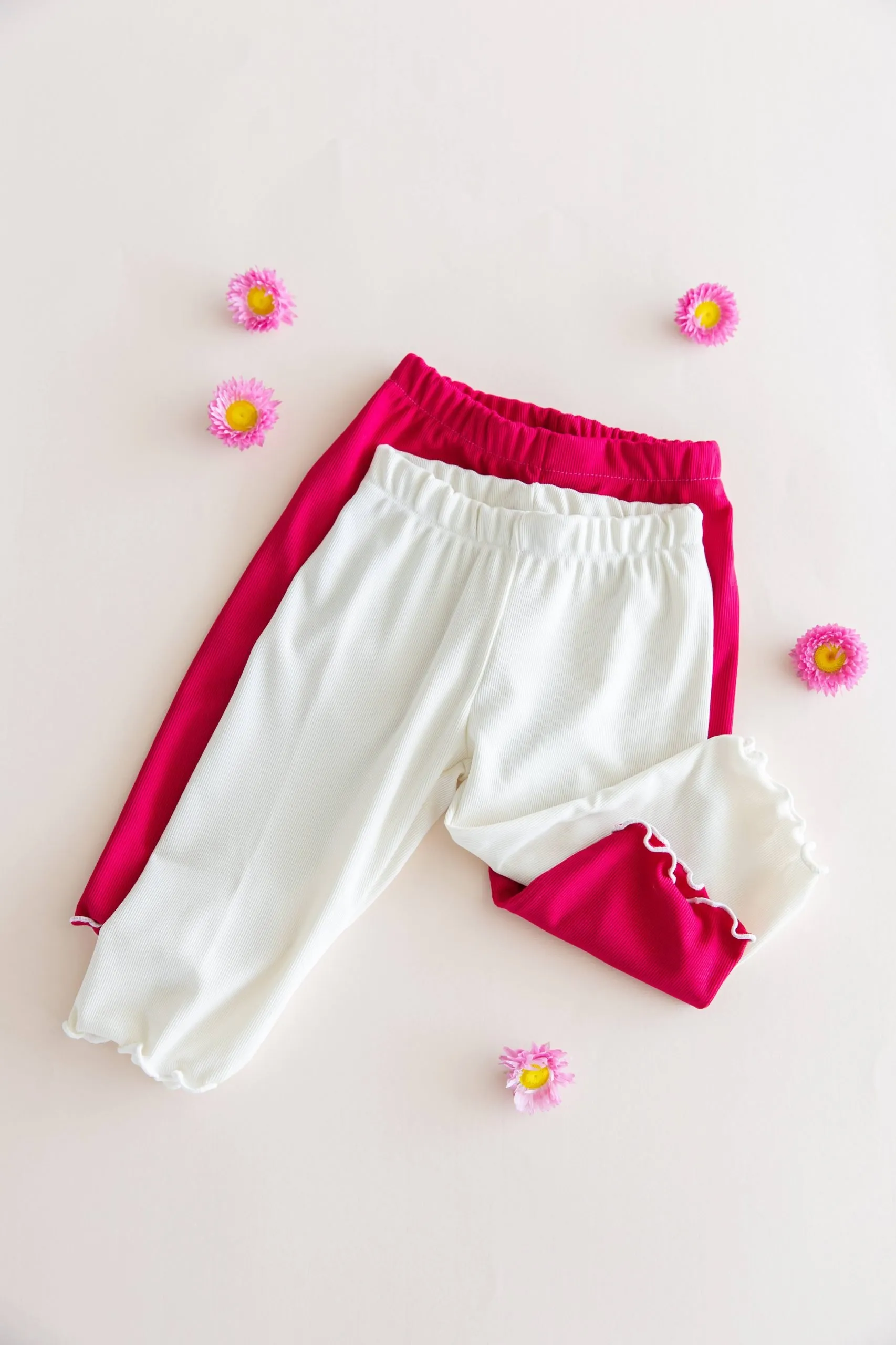 Dos calzas ciclistas infantiles de tela morley, color blanco y color fucsia magenta, mostrando el detalle del ruedo con terminación ruloté.