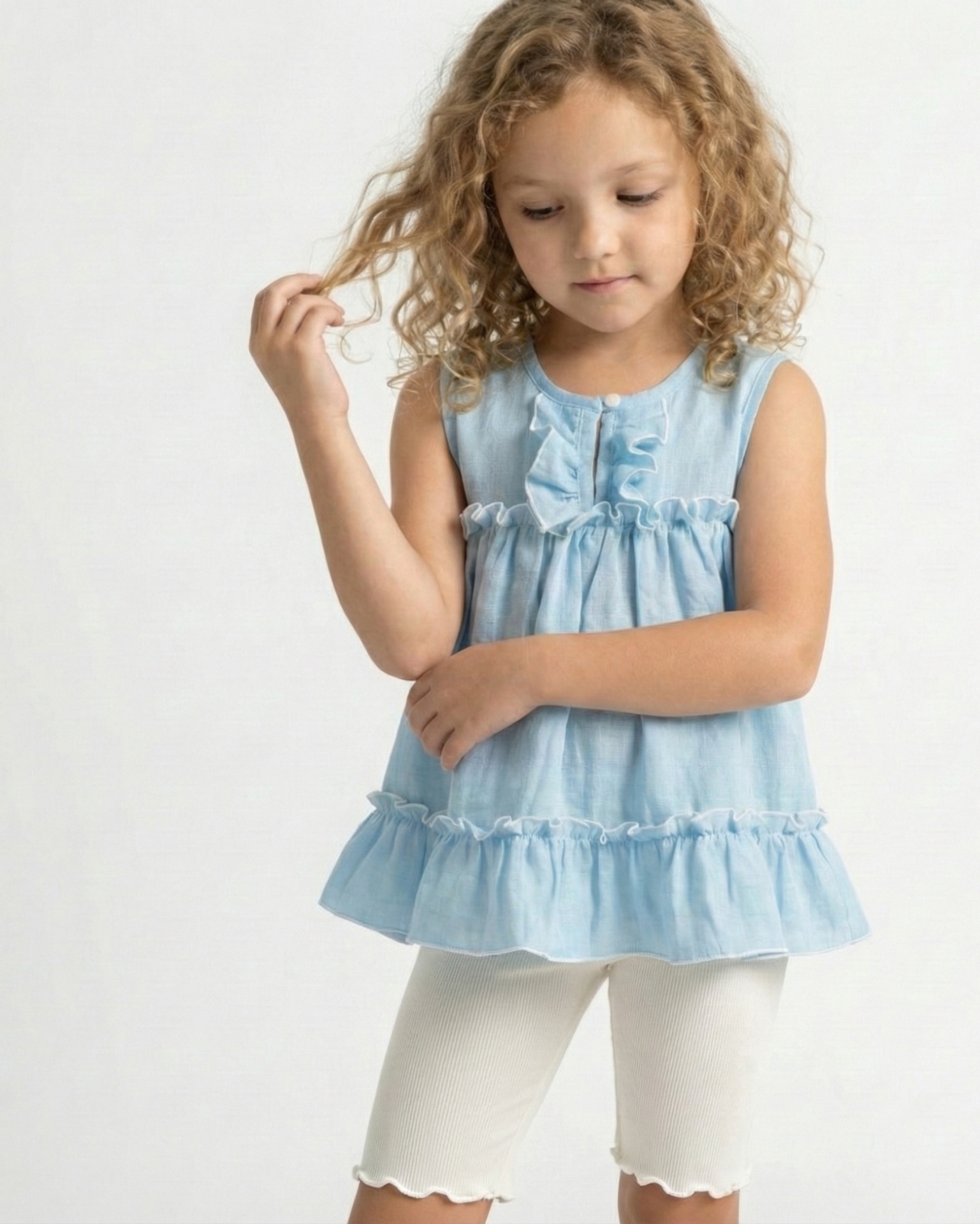 Niña con cabello rizado usando una blusa celeste sin mangas de tela gasa tipo lino, con volados y bordes blancos festoneados. Ropa de verano elegante para niñas.