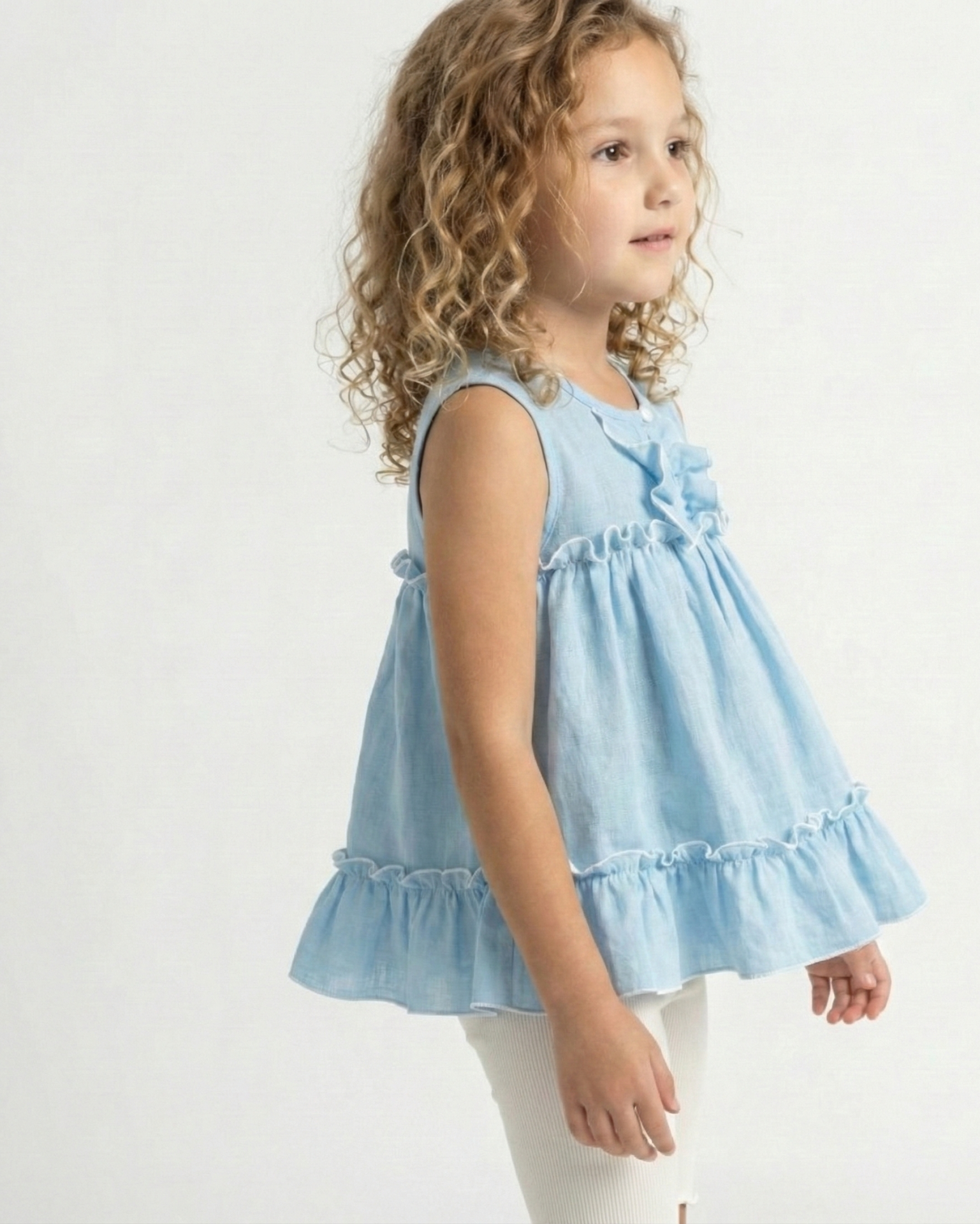 Niña con cabello rizado usando una blusa celeste sin mangas de tela gasa tipo lino, con volados y bordes blancos festoneados. Ropa de verano elegante para niñas.