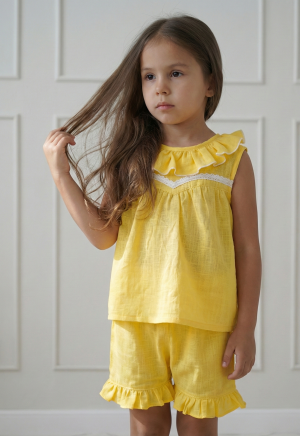 Conjunto de dos piezas para niña color amarillo maíz extendido sobre fondo rosa con flores. Incluye blusa con volados y puntilla blanca, y pantalón tipo bombachudo amplio de tela gasa texturada.