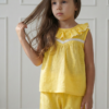 Conjunto de dos piezas para niña color amarillo maíz extendido sobre fondo rosa con flores. Incluye blusa con volados y puntilla blanca, y pantalón tipo bombachudo amplio de tela gasa texturada.