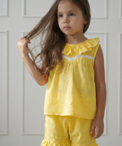 Conjunto de dos piezas para niña color amarillo maíz extendido sobre fondo rosa con flores. Incluye blusa con volados y puntilla blanca, y pantalón tipo bombachudo amplio de tela gasa texturada.